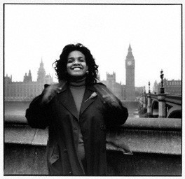 Diane Abbott in 1987, with the Houses of Parliament in the background. She is a black woman, smiling and waving her hands in excitement.