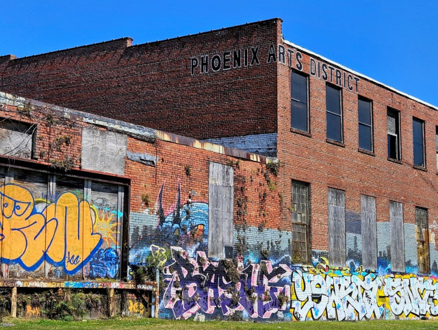 Large, two story, red brick warehouse long abandoned. Boarded up windows and doors. Dark ivy like vines climb the exterior walls. The top corner has Phoenix Arts District painted in large black letters. The ground level building and delivery dock were once decorated with murals of local Jacksonville scenes, now nearly completely covered in large bubble letter graffiti.