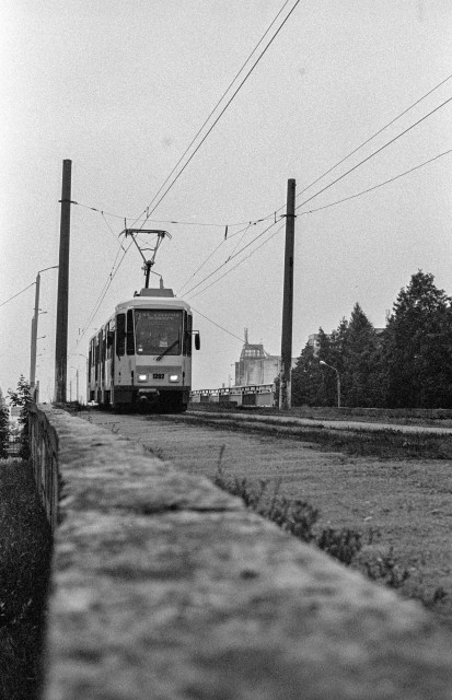 The image is a black and white photograph of a tram moving along a track. The tram, labeled with the number "1202," is centered in the frame, and the scene has a vintage, grainy quality. Overhead power lines and poles are visible, indicating the electric nature of the tram. The background features a mix of trees and buildings, with one notable building having a distinct, somewhat industrial or historical architectural style. The foreground includes a stone or concrete barrier that runs parallel to the tram tracks. The overall atmosphere of the photograph suggests an urban or suburban setting