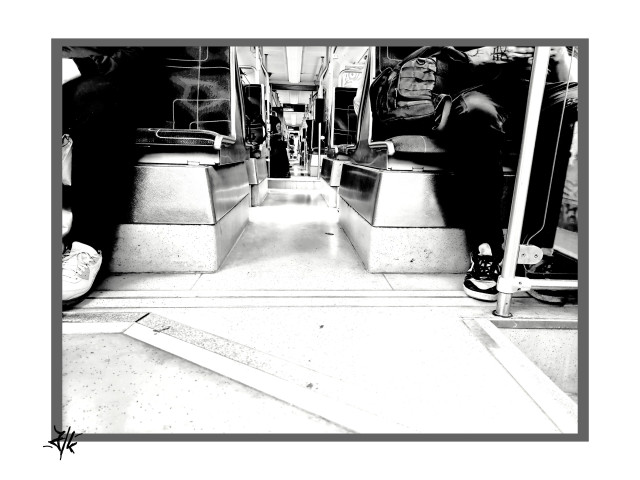 Schwarz-Weiß Foto aus der Straßenbahn. Die Perspektive ist aus Fußboden Nähe, Mitte vom Gang. Zu sehen sind die Sitze mit den Beinen der Fahrgäste.