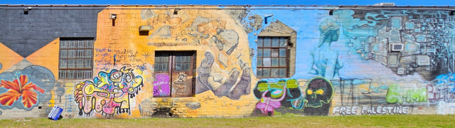 Old abandoned warehouse painted with a variety of art, murals, and graffiti.  This section, a wide view, including a human hand reaching out from a warehouse window; a Mardi-Gras style trumpet player; a cartoon gorilla and day-of-the-dead skull; a nude woman overlooking a desert, a large one-eyed 'Alien' style creature guardian an endless mountain range of old computer monitors all wired to the creature; a cartoon dragon.