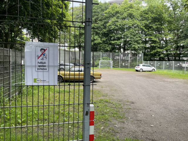 Ein eingezäunter Bolzplatz. Am Eingang hängt ein „Fußball-spielen verboten“ Schild. Auf dem Bolzplatz parken Autos. 