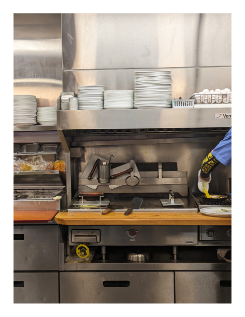 food prep line in a waffle house, seen from the counter. an unseen cook's arm peeks in at right wearing a blue short-sleeve shirt over long-sleeve tee with the WH logo in yellow and black, he's pouring scrambled eggs from a small bowl into a pan.