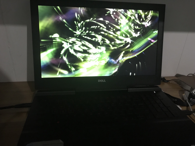 A Dell Inspiron 15 7567 Gaming laptop with a screensaver running. The screensaver has lots of colourful curves: green, white, purple. The image is not very good because the bright light has bloomed in the camera.
