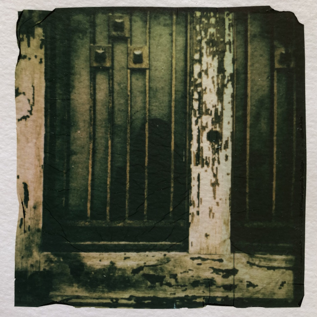 A door with windows and bars, like a prison, and the reflection of a black silhouette in the window. Polaroid emulsion lift. 
