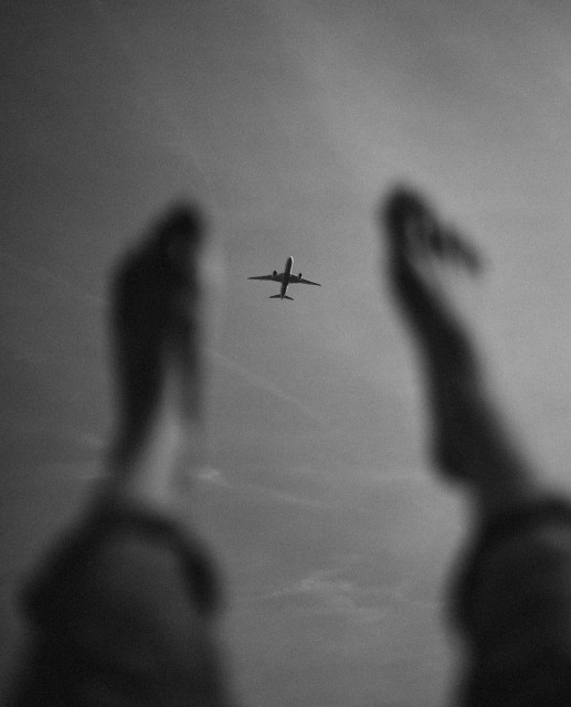 Photography. A black and white photo of two blurred bare feet and an airplane. The focus is on a small airplane that can be seen in the sky between the two outstretched legs. As if you were lying in the grass on a sunny day, looking up at the sky, and not even an airplane could disturb you.
