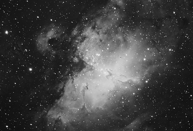 A bright deep space nebula in black and white. A cluste rof bright stars sits in the middle, surrounded by softly glowing gas. Finger-like pillars of gas and dust point towards the middle. These are the pillars of creation and inside them stars are being formed.