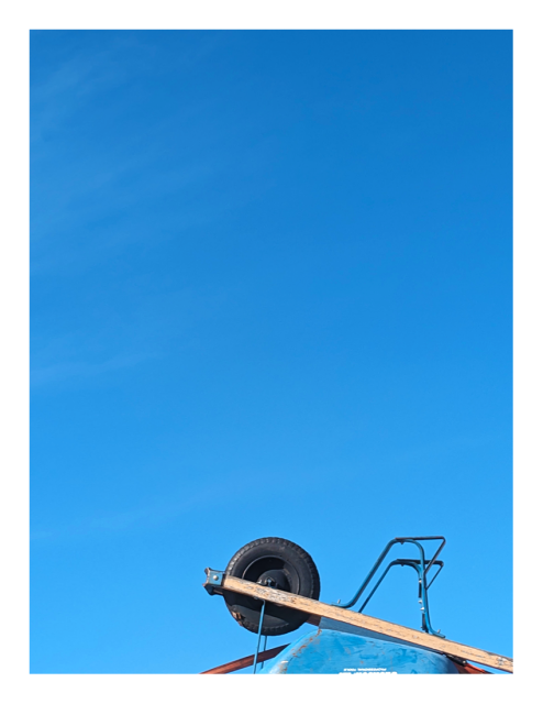 daytime. a wheelbarrow with blue bucket and wood handles is upside-down and strapped to the roof of an unseen truck. the background is a matching blue sky.