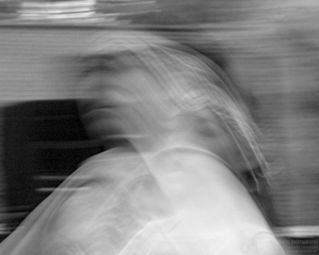 This is a long exposure in black and white of a powwow dancer. You can see two ghostly faces that swoosh from right center to upper left. It's as though the dancer jumped and turned their head to their right during the exposure. A zig-zag beaded headband can be discerned from the photo. Everything else is a blur. 