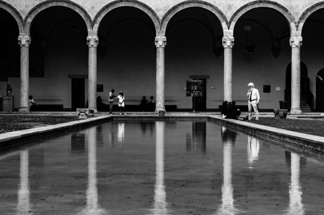 Fotografía en blanco y negro de uno de los patios del palacio de los Sforza en Milán (Italia) donde se ve un estanque en el cual se reflejan unos arcos además de algunos visitantes.