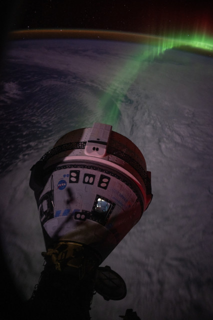 Boeing Starliner in the foreground, while Earth is in the background with a line of vivid green aurora visible