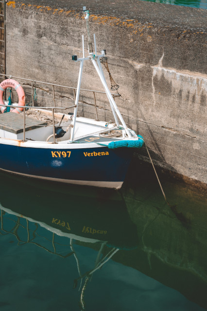 A photo of a boat in a harbour. The framing of the photo is so that you see the front half of the board, the barbour wall, and the reflection of the boat in the slightly rippled water. Otherwise the frame is free of the usual harbour clutter.