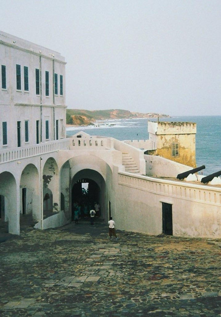 Point of no return: Ghana. Crucial point in African human trafficking; once you past this point, you will never return to the continent in your lifetime. A historic white stone building with arches and cannons overlooking a coastline. Several people walking under an archway.