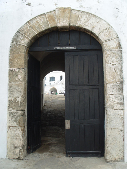 A stone archway with a partially open black wooden door, labeled "Door of Return," leading to a cobbled path and a white building in the background.