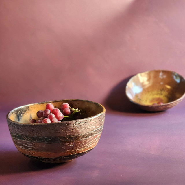 Two handmade bowls, striped in deep purple, black, and leather tones with carved geometric designs. The interior are shades of brown and gold flecks. The background is aubergine.