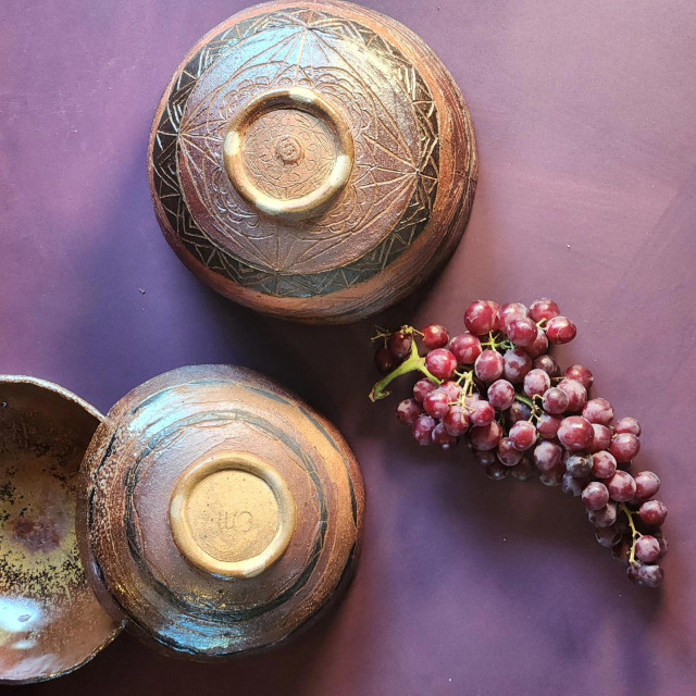 Three inverted handmade bowls to show the exterior carving on bands of deep red/purple, black, and leather color. The background is aubergine and there are grapes in the foreground.