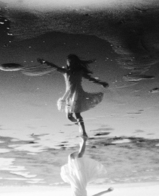 Photography. A black and white photo of a girl dancing on the beach. A young woman in a wide white dress dances in the puddles on the beach with her arms outstretched. She is reflected in the water. But it looks as if she is dancing through the wind in a sky full of clouds. The special thing about it is that the photo is actually upside down.  If you turn the photo around, you can see her entire reflection in the water, but at the top you can only see her up to her neck.  The unusual perspective makes the photo appear strange, we are irritated and look for the artistic photographic alienations. But it is simply upside down.