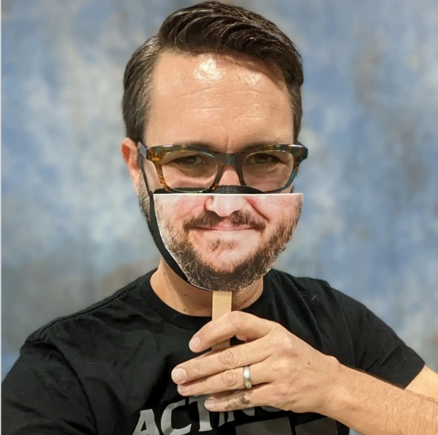 Wil Wheaton is wearing horn-rimmed glasses, a black respirator and is holding up a cardboard cutout of the lower half of his face on a tongue depressor up over his respirator.