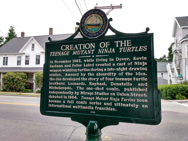 A green heavy duty, serious historical marker sign with the seal of New Hampshire at the top, with a charming clipper ship looking vessel on the water, surrounded by state incorporation date stuff. 

Text of the sign: 
CREATON OF THE TEENAGE MUTANT NINJA TURTLES

In November 1983, living in Dover, Kevin Eastman and Peter Laird created a cast of Ninja weapon-wielding turtles during a late-night drawing session. Amused by the absurdity of the idea, the duo developed the story of four teenage turtle brothers: Leonardo, Raphael, Donatello and Michelangelo. The one-shot comic, published independently by Mirage Studios on Union Street.  Teenage Mutant Ninja Turtles soon became a full comic series and ultimately an international multimedia franchise. 

2023