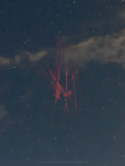 Photograph of a red sprite. It has three vertical columns that branch out towards the top and and in the middle. The thing looks a bit like someone's crude attempts to paint lines with a can of spray paint. The color is a very vibrant red against a blueish nightsky with many stars. 