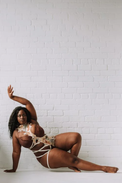 A black woman in brown lacy underwear wrapped with cream ribbons and adorned with pastel flowers. She's stretched out, lifting herself up on one arm and one leg, the other arm stretched up over her head, fingers reaching upwards. She's looking straight at the camera, evocatively. She's posed against a white brick wall on a white floor.