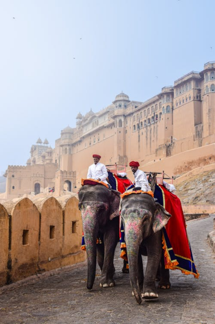 Two men riding elephants. The elephants have colourful patterns painted onto their trunks and ears, and are draped with red, blue, and gold cloths. The men wear white shirts and red hats. A wall of sandstone arched shapes lines the roadside, and in the distance, a towering city wall stretches away against a grey sky, with rows of windows at the top and domed roofs just visible through the grey.