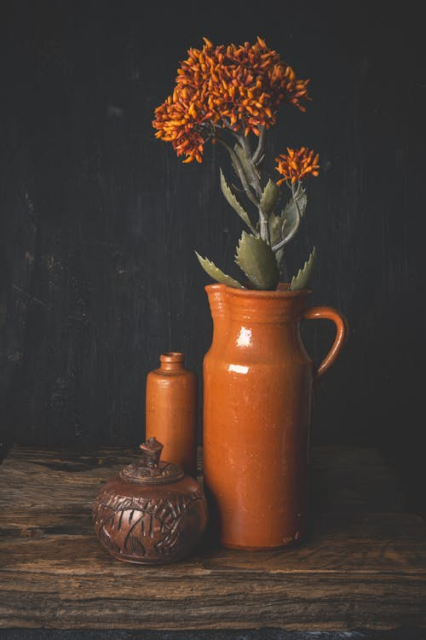 A still life against a dark wooden surface of a small round carved wooden bowl and lid, a dark orange glazed tall narrow vase, and a similarly glazed tall orange jug with a ridged lip. There are orange flowers with green spiky leaves in the jug.