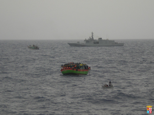 A picture of an overcrowded fishing boat with refugees, in the background a larger warship.

http://static-cdn.sr.se/images/4763/3465846_2048_1152.jpg?preset=1024x576