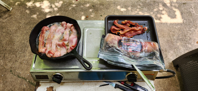 An outside gas stove with bacon cooking in an iron skillet on the left and some uncooked and cooked bacon on the right.