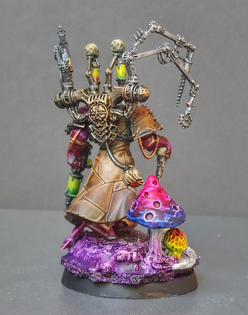 Painted miniature of Fabius Bile. He is walking over a rocky base (mostly painted in purple tones, but some are green/brown, too) with giant mushrooms and skulls. He is holding Torment and some kind of Injector. Back View. the Chirurgeon is very worn-metallic looking.