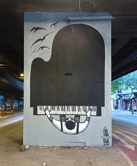Streetartwall. On a gray concrete pillar under a street bridge in the city center, a large black and white mural of a piano player was sprayed/painted in a simple comic style. The background is gray. The whole thing is depicted with a view from above of a large piano on which a dark-haired man is playing the keys with both hands. A flock of black birds flies past at the top of the picture. Streets can be seen to the right and left of the mural. The unusual perspective, the minimalist drawing and the huge piano make the mural so special.