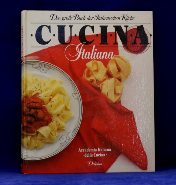 A white hardcover book

On a red table cloth, a plate with tagliatelle and tomato sauce.

On the table, beside the plate, a few dry noodles and a glass of red wine