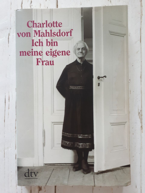 Cover of the book "Charlotte von Mahlsdorf. Ich bin meine eigene Frau". A page-sized photo of Charlotte in a long black dress. Charlotte is standing in a doorway with the arms crossed behind the body. The photo was probably taken in Charlotte's own old manor house (now the Gründerzeitmuseum).