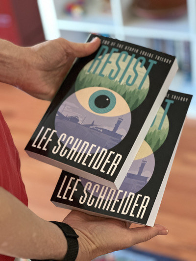 Two hands holding a novel called Resist by Lee Schneider. The cover art features an eye in the middle. 