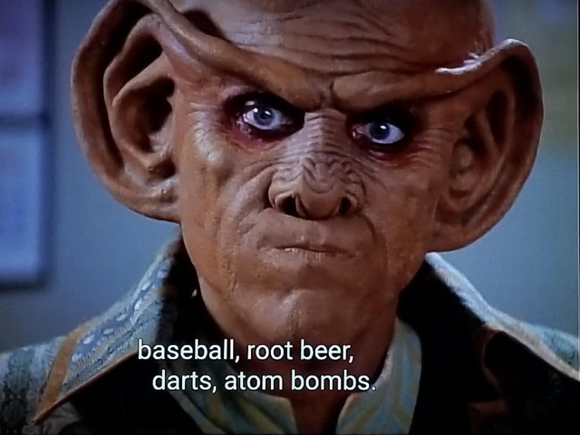 Close up on Quark, a Ferengi with a smooth domed head and ears that are giant and ridgey and a nose that kind of looks like a pine cone.  He is pursing his mouth and doing a Jim from the office "oh brother" look right at us. Closed caption reads, "baseball, root beer, darts, atom bombs."