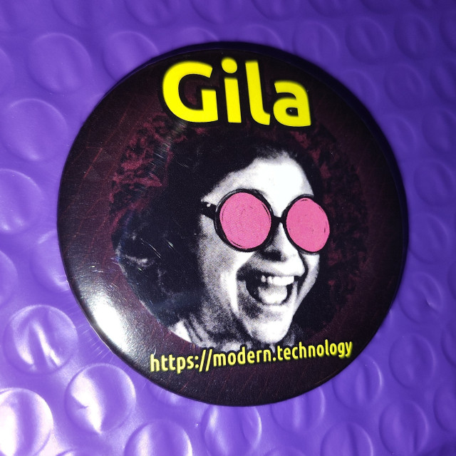 Photo of a button badge with a stylized photo of Gila, captioned with her name and the URL https://modern.technology (which is where she does podcasting with me.) The featureless pink cartoon eyes of the Modern Technology Podcast Network mascot have been crudely drawn onto her face, which is a design conceit done to all photos of people on the Modem Technology Podcast Network site for some reason.