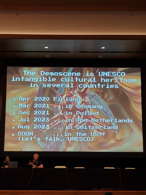 A photo of two presenter's and a big screen showing a timeline of Demoscene being declared UNESCO intangible cultural heritage in different countries. Apr. 2020 Finland, Mar 2021 Germany, Dec 2021 Poland, Jul 2023 The Netherlands, Aug 2023 Switzerland.