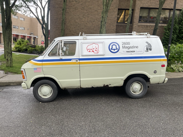 The 2600 van parked on a curb with its Bell Atlantic logo. It’s green half way from the bottom, with a yellow and blue stripe going through the middle of the van. The top half of the van is white with a HOPE logo and a 2600 magazine lettering. 