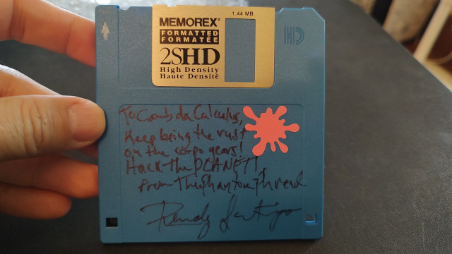 A blue floppy disk; there is a hand written autograph on the label area that reads:

"To LambdaCalculus,

Keep being the rust on the corpo gears! Hack the PLANET!
From The Phantom Phreak,
Remoly Santiago"

A pink splotch sticker is affixed to the upper right corner
