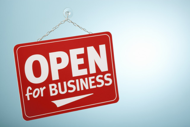 A red sign with white lettering that reads “Open for business” hanging by a golden chain from a hook attached to what appears to be frosted glass by a plastic sucker.