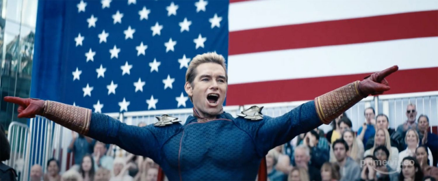 A scene from The Boys series, Homelander pointing at crowd with a huge USA flag behind him.