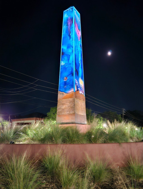 An obelisk stands tall above a decorative roundabout beneath dark night skies. Ground level lighting adds to the bright colored sculpture and it's glow of blue sea scenes.