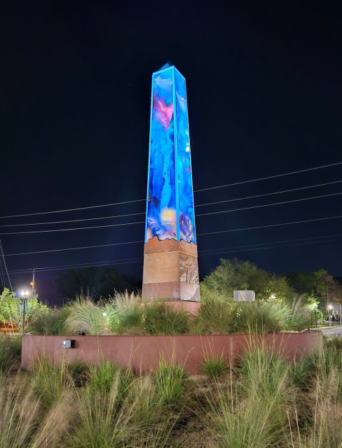 An obelisk stands tall above a decorative roundabout beneath dark night skies. Ground level lighting adds to the bright colored sculpture and it's glow of blue sea scenes.