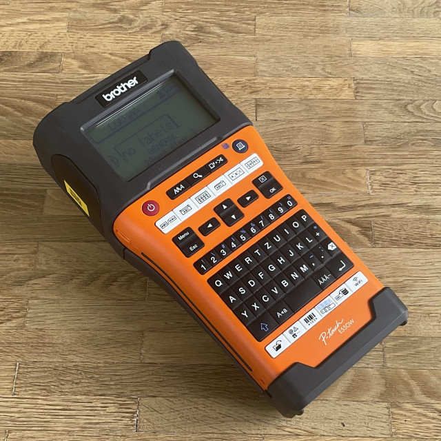 Photo of a Brother ptouch e550w Labelwriter on a wood floor. The device looks quite large and heavy, like a tool, and half of it is orange so it does not get lost easily.