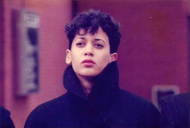 A young Kamala Harris, wearing a black coat with a high collar in front of a blurred maroon background. She has short hair and kind of a wistful look on her unsmiling gorgeous face. She about to drop the most fire synth album ever.