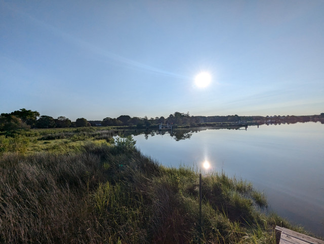 Photo of a clear morning sunrise over Harris Creek sun rising low to the left in a cloudless blue sky. Marsh is green and ready for the day. Creek water is calm and perfectly reflects the sun and sky.