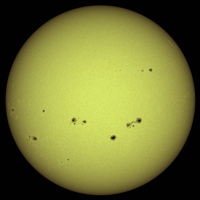 A yellow sun with multiple dark spots, known as sunspots, scattered across its surface. The background is black.