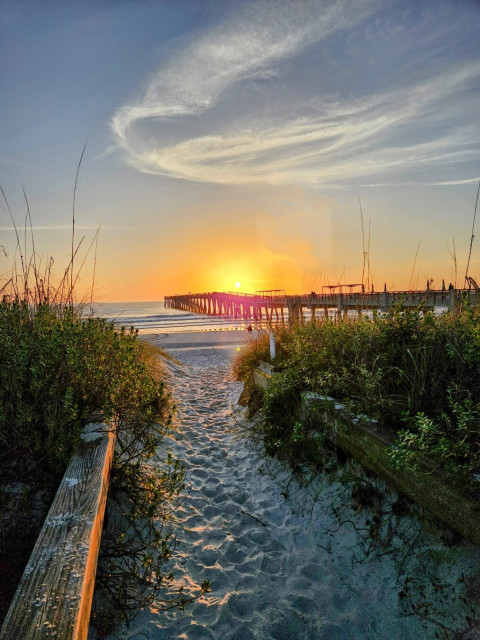 From the white sand dunes approaching a beach on the Atlantic coastline during sunrise.  With a thick layer of white sand in a pathway between green brush, a bright yellow sun rises over a long wooden pier extending far out into the ocean's surf, beneath sunny, bright blue skies, casting shades of yellow and orange across the horizon.