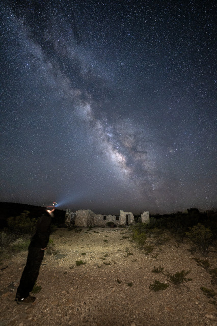 A man standing at the ruins of an old ghost town with his headlamp shining off towards the Milky Way