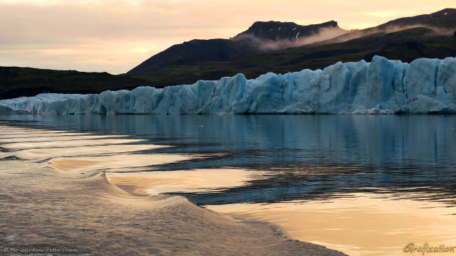 A photo of a landscape, with a foreground of water and a horizon line of low mountains. The sky is pastel orange and yellow; it's near to sunset. This colour is reflected in the foreground water surface. At the centre of the shot is a wide line of tall cyan ice slabs, arranged in a row. This is the glacier snout, with crevasses visible as cracks. Above that is the dark rock face of its containing mountain. The water surface is being disturbed by the movement of the boat in which the viewer is seated. Ripples and interference patterns are creating a beautiful abstract of orange and cyan triangles, picking up both the ice and sky colours.